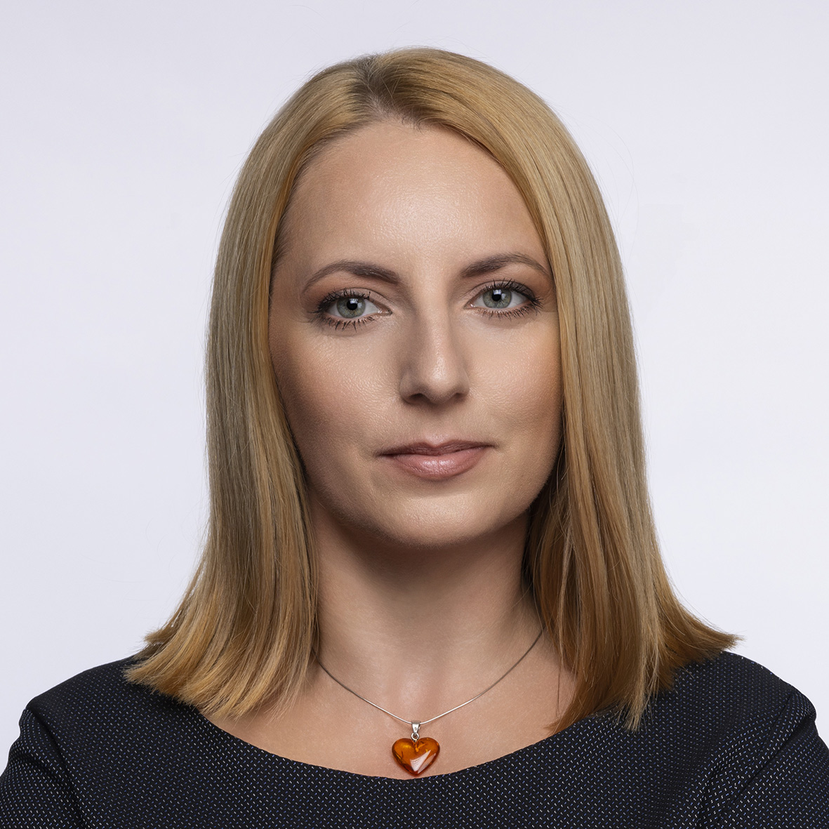 Ewelina Bugajska, z blond włosami sięgającymi ramion, w ciemnym topie i z naszyjnikiem z wisiorkiem w kształcie serca. Stoi na prostym tle, patrząc prosto w kamerę.