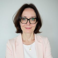 Agnieszka Pipała, kobieta z brązowymi włosami sięgającymi do brody i okularami, uśmiecha się delikatnie. Ma na sobie bladoróżową marynarkę na białym topie, ozdobioną złotym naszyjnikiem i kolczykami, pozuje na jasnym tle.