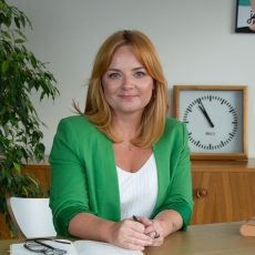 Anna Stępkowska, kobieta o blond włosach w zielonym blezerze, siedzi przy biurku, trzyma długopis i się uśmiecha. W tle widać notatnik, okulary, roślinę i zegar ścienny wskazujący 3:15.