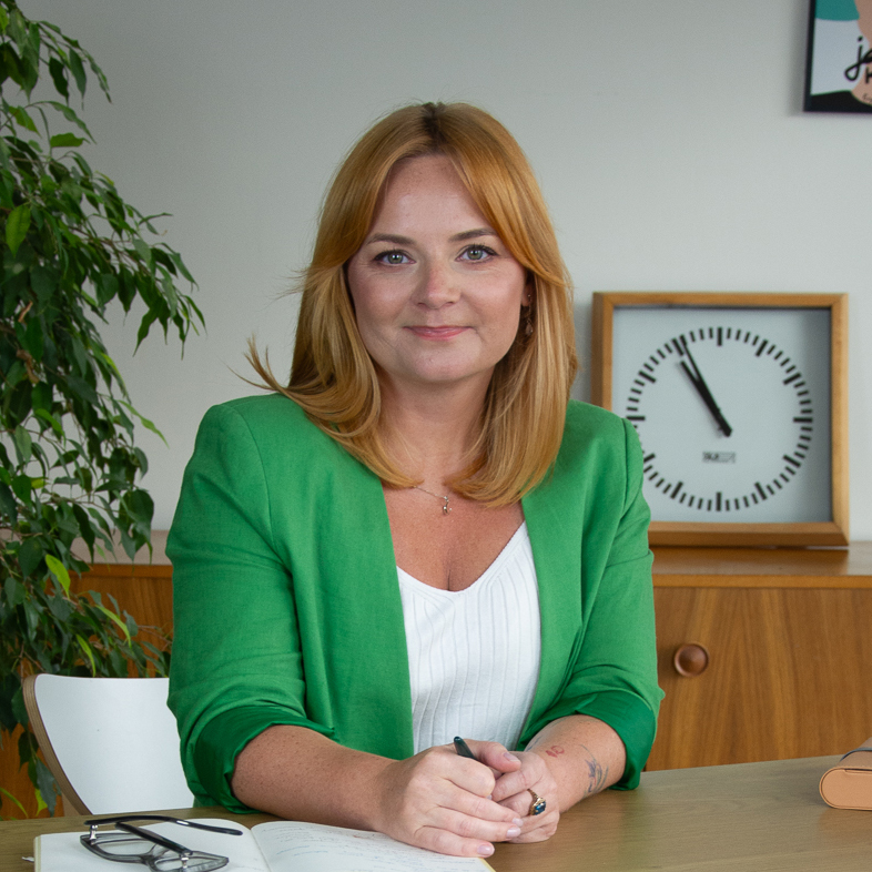 Anna Stępkowska, kobieta o blond włosach w zielonym blezerze, siedzi przy biurku, trzyma długopis i się uśmiecha. W tle widać notatnik, okulary, roślinę i zegar ścienny wskazujący 3:15.