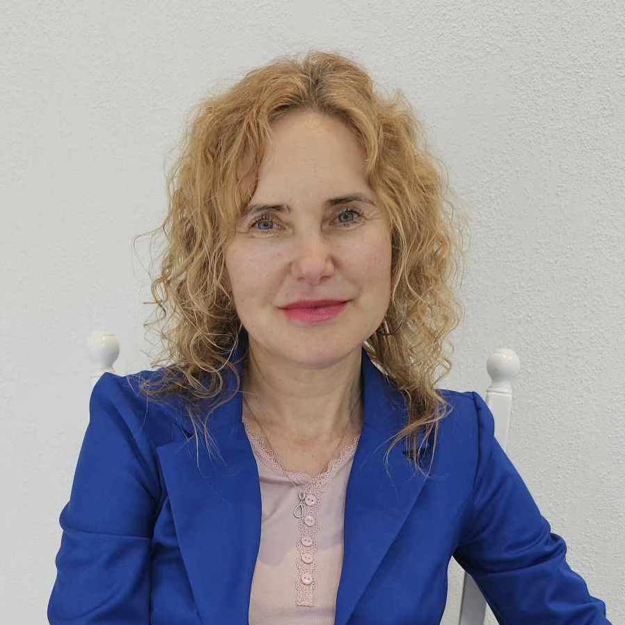 Agnieszka Kaźmierska, kobieta o kręconych blond włosach, uśmiecha się, siedząc na jasnym tle. Ma na sobie jasnoniebieską marynarkę na różowej bluzce, emanując pewnością siebie i stylem.