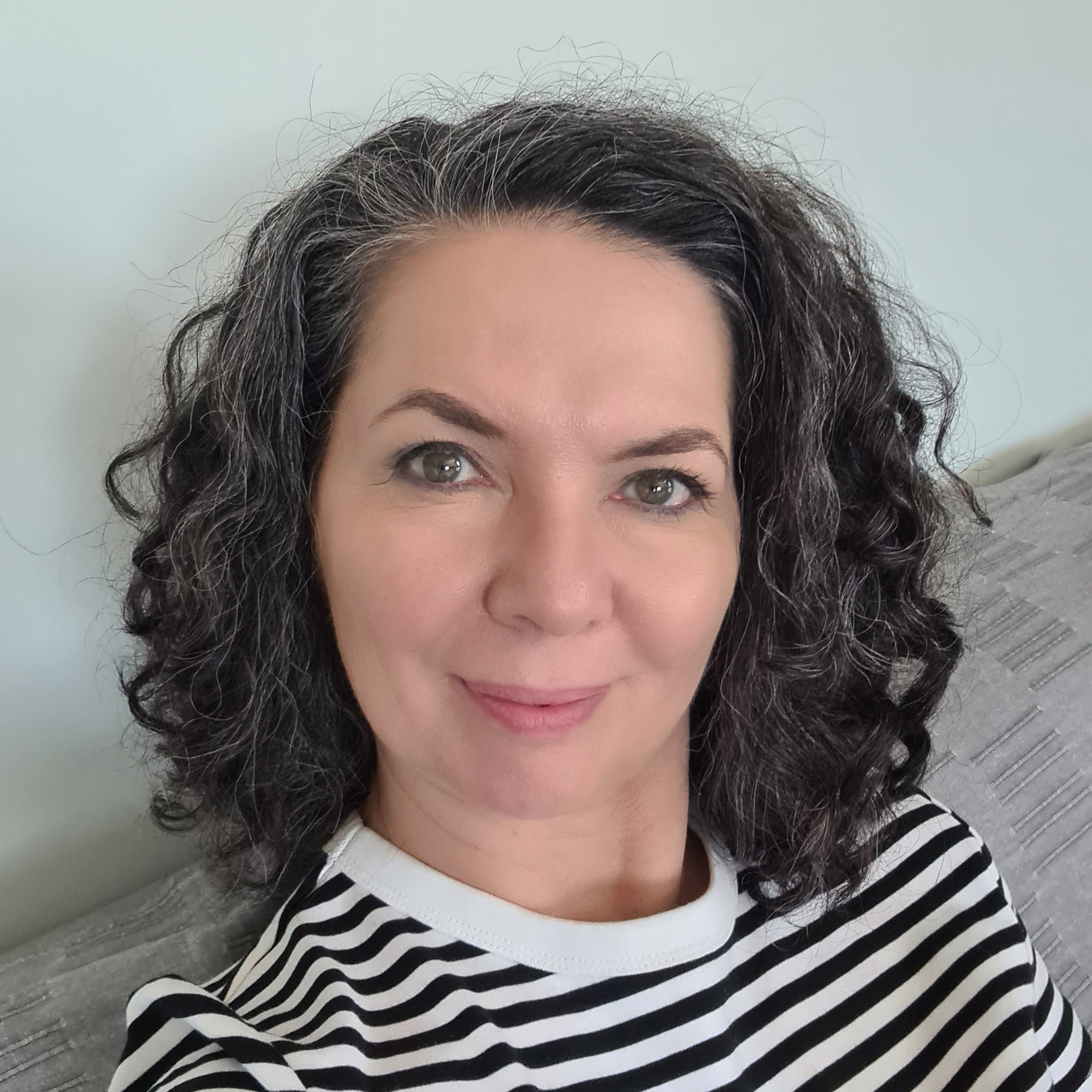 Anna Miądowicz-Ułamek, kobieta o kręconych, ciemnych włosach z siwymi refleksami, pozuje do selfie. Ma na sobie czarno-białą koszulę w paski i delikatnie uśmiecha się do kamery. W tle znajduje się neutralna sofa.