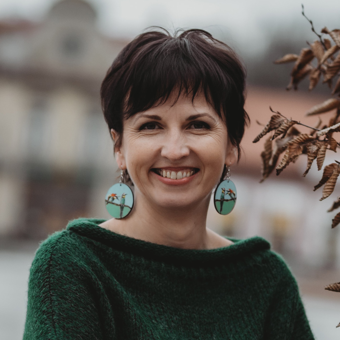 Anna Wólczyńska-Cholewa, kobieta o krótkich ciemnych włosach i turkusowych kolczykach, uśmiecha się, stojąc na zewnątrz. Ma na sobie zielony sweter, a w tle widać rozmazane budynki, a na pierwszym planie suche liście.