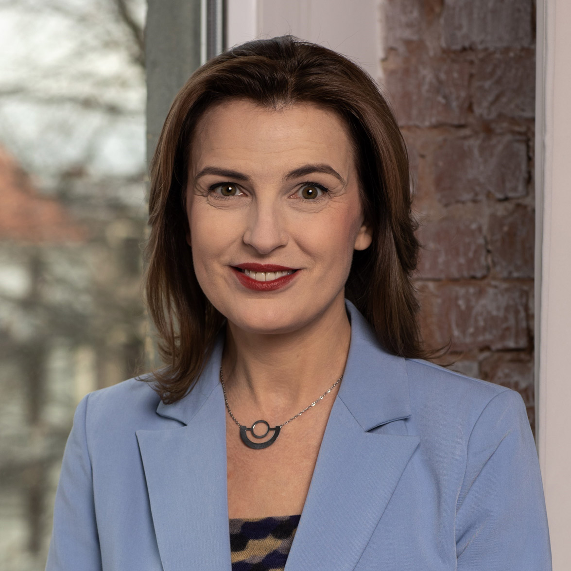 Ewa Stępniewicz, kobieta z brązowymi włosami sięgającymi ramion, nosi niebieski blezer i wzorzysty top. Uśmiecha się ciepło przed oknem, a rozmyte tło ceglanej ściany i drzew dodaje spokojnej atmosfery.
