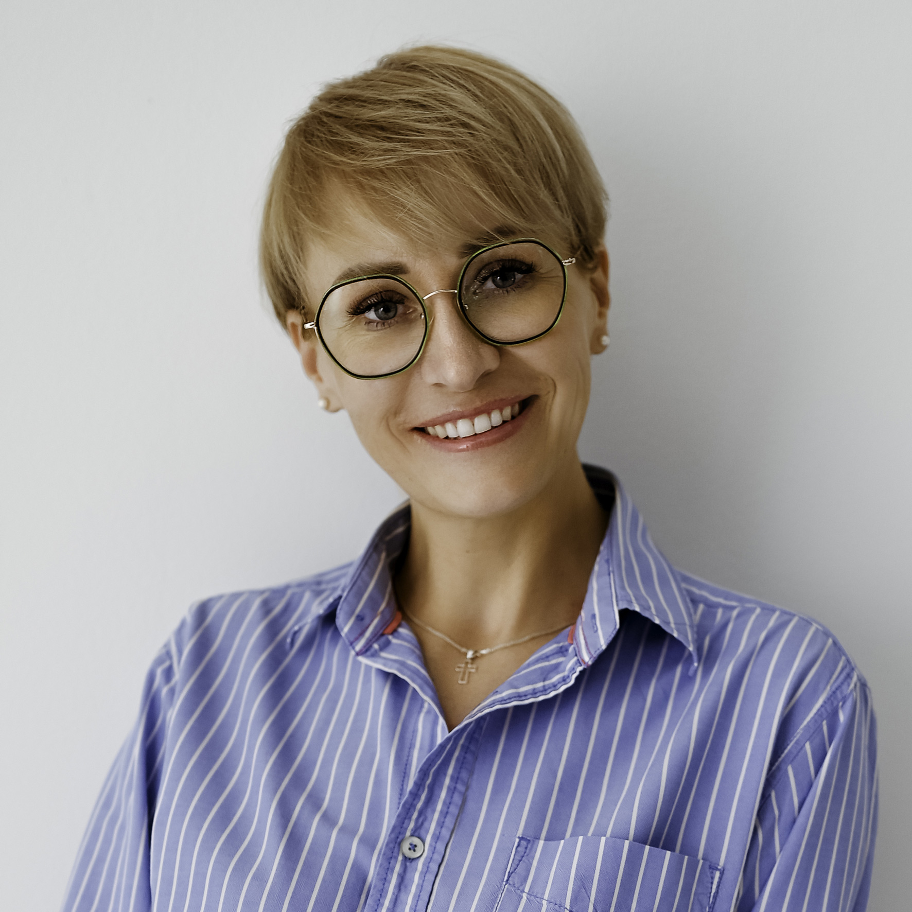 Aleksandra Szary, z krótkimi włosami i w okularach, uśmiecha się serdecznie, mając na sobie koszulę w niebiesko-białe paski na jednolitym tle.