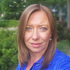 Sylwia Zakrzewska, kobieta o prostych, jasnobrązowych włosach i niebieskich oczach, lekko uśmiecha się do kamery. Ma na sobie królewski niebieski top i złote kolczyki. Tło jest na zewnątrz, z zielonymi liśćmi i drzewami nieostrymi.