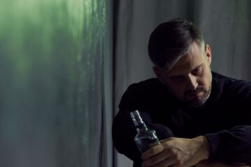Mężczyzna z krótkimi włosami siedzi w słabo oświetlonym pokoju, wygląda na zamyślonego. Trzyma pustą szklaną butelkę, sugerując walkę z uzależnieniem od alkoholu i opiera się o teksturowaną, zielonkawą ścianę. Jego wyraz twarzy jest zamyślony i nieco ponury.