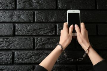 Ręce w kajdankach trzymają smartfon przy czarnej ścianie z cegieł, symbolizując walkę z fonoholizmem o uwolnienie się od uzależnienia od telefonu.