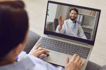 Osoba siedząca na kanapie używa laptopa do rozmowy wideo skupionej na terapii uzależnień. Lekarz w białym fartuchu uśmiecha się i gestykuluje otwartą dłonią z nowoczesnego biura. Na ekranie laptopa wyświetlany jest interfejs rozmowy.
