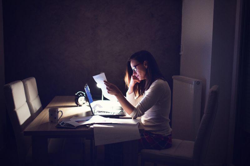 Kobieta siedzi przy stole późną nocą, pracując na laptopie z papierami w ręku. Wydaje się skupiona, ucieleśniająca pracoholizm, z kubkiem, kalkulatorem i zegarem obok siebie, oświetlonymi światłem komputera.