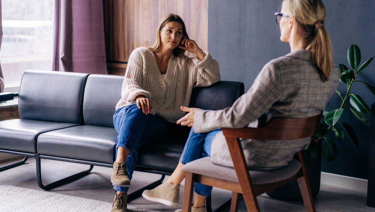 Dwie kobiety siedzą naprzeciwko siebie w nowoczesnym pokoju, rozmawiając o co to jest psychoterapia. Jedna kobieta słucha uważnie z kanapy, podczas gdy druga gestykuluje, wyjaśniając na czym polega psychoterapia.
