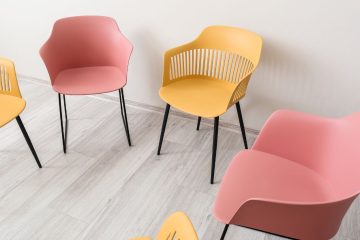 Cztery nowoczesne krzesła z czarnymi metalowymi nogami, dwa różowe i dwa żółte, ustawione są w półkolu na jasnej drewnianej podłodze przy białej ścianie – to przyjazna przestrzeń, idealna do sesji psychoterapii.