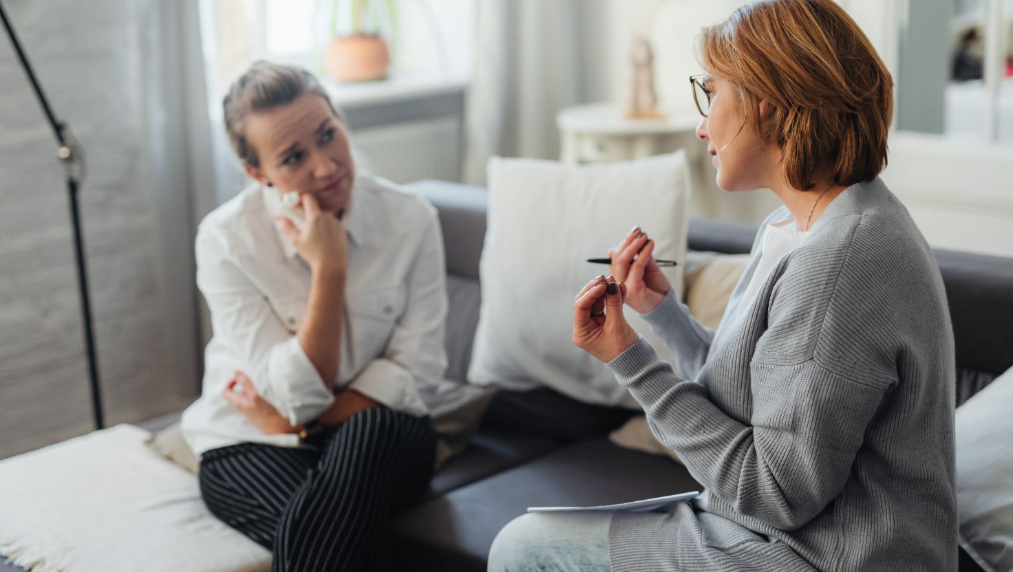 Dwie kobiety siedzą na kanapie w salonie i rozmawiają – jedna słucha w zamyśleniu, druga gestykuluje długopisem. Spotkanie wygląda na sesję psychoterapeutyczną.