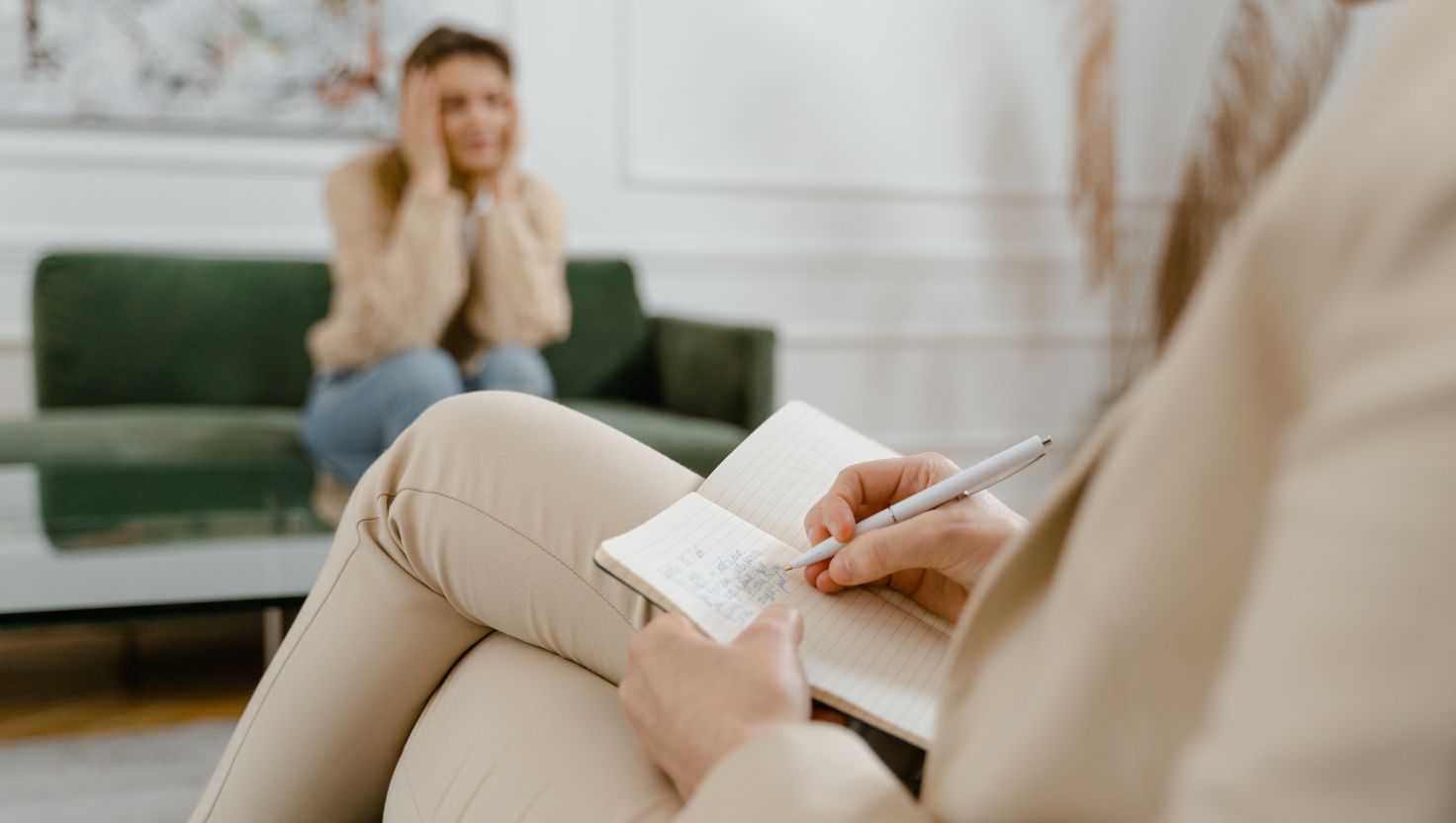 Osoba siedzi na zielonej kanapie z głową w dłoniach, wyglądając na przygnębioną, podczas gdy inna robi notatki w notatniku podczas sesji psychoterapii integracyjnej, ilustrując na czym polega nurt integracyjny w terapii.