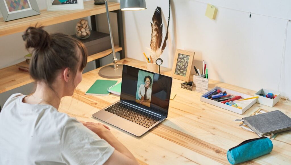 Osoba siedzi przy drewnianym biurku, oglądając uczestnicząc w sesji terapii psychodynamicznej online. Na biurku znajdują się artykuły piśmiennicze, notatniki, lampa i przybory artystyczne, tworząc przytulne miejsce do pracy.