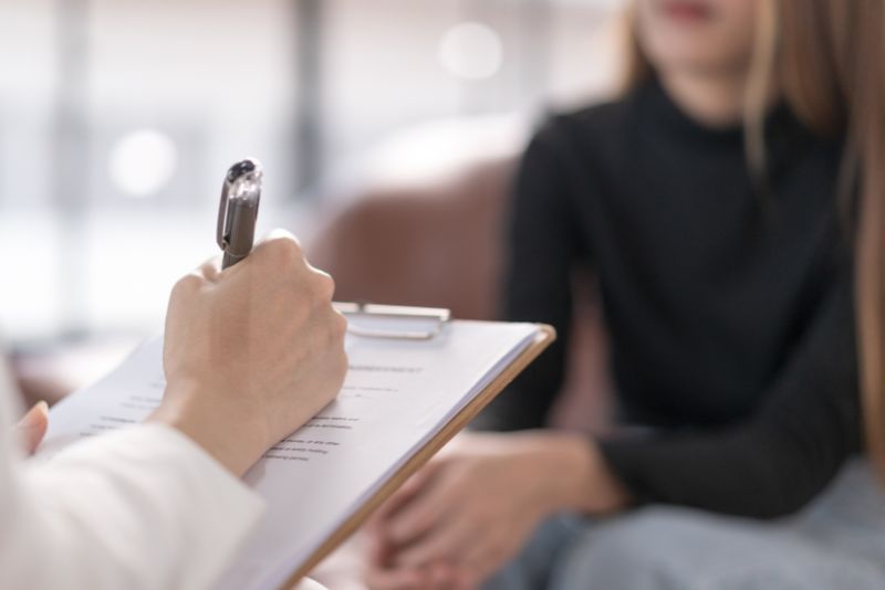 Osoba trzymająca schowek i długopis wydaje się robić notatki, podczas gdy inna siedzi naprzeciwko niej, co sugeruje sesję doradczą skoncentrowaną na psychosomatyce lub problemach emocjonalnych. Twarz jest nieostra.