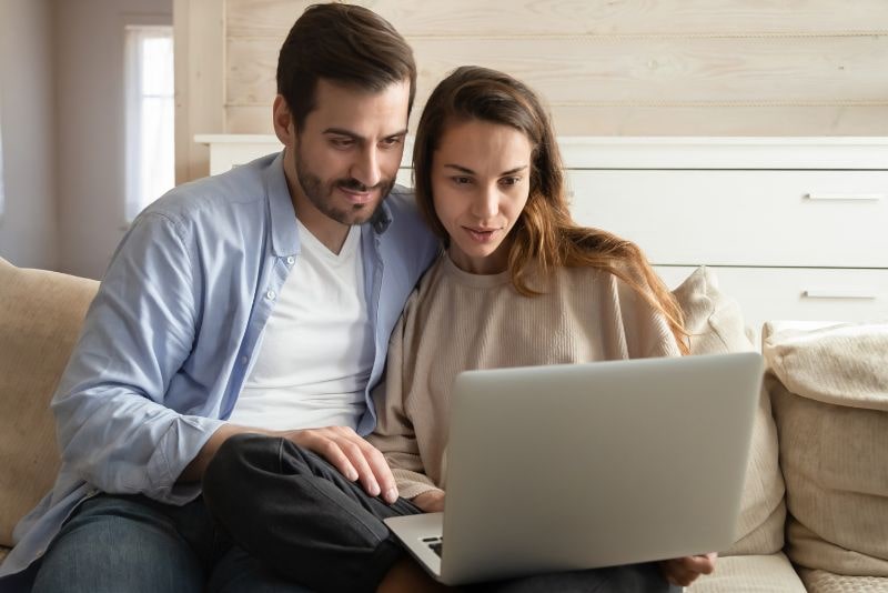 Para siedzi blisko siebie na kanapie, patrząc z zainteresowaniem na ekran laptopa. Zaangażowani i skupieni, uczestniczą w psychoterapii par, podejmując proaktywne kroki w kierunku lepszego zdrowia psychicznego razem.