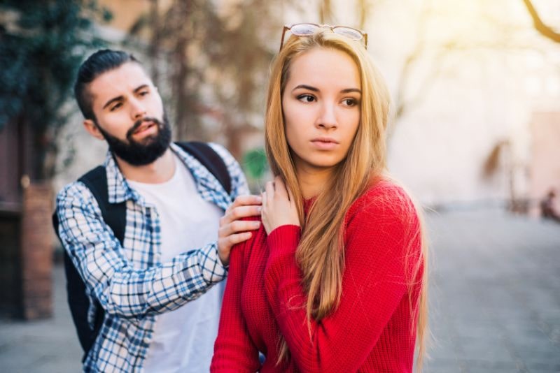 Mężczyzna z brodą delikatnie trzyma kobietę za ramię, podczas gdy ona odwraca wzrok z poważnym wyrazem twarzy, zastanawiając się nad tym, jak sobie poradzić podczas kryzysu w związku. Są na zewnątrz w słoneczny dzień, a kobieta wydaje się zdenerwowana lub zdystansowana.