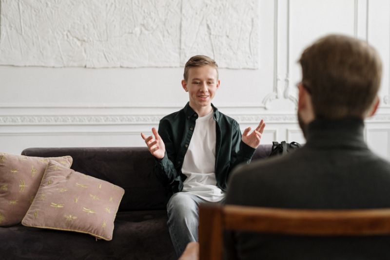 Młody mężczyzna siedzi uśmiechnięty na kanapie i opowiada terapeucie o swoich emocjach oraz odczuciach. Jego uśmiech i gestykulacja mogą świadczyć o pozytywnym wpływie terapii.
