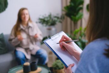 Osoba trzymająca notes i długopis robi notatki, podczas gdy inna siedzi na kanapie i opowiada o odczuwanych emocjach, co sugeruje sesję terapeutyczną.