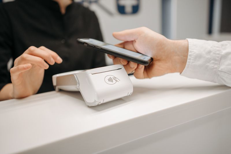Osoba przykłada telefon do białego terminala płatniczego, aby uregulować należność za umówioną sesję terapeutyczną.