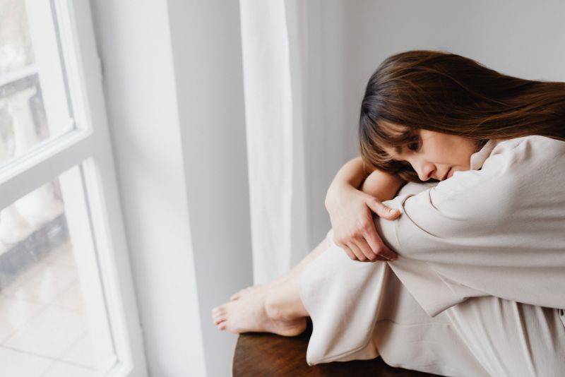 Kobieta siedzi przy oknie, obejmując swoje nogi i przyjmując pozycję obronną, co jest efektem odczuwania smutku, który jest spowodowany wyuczoną bezradnością.