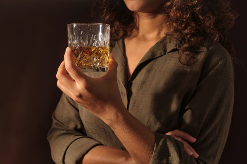 Kobieta z kręconymi włosami, ubrana w oliwkowozieloną koszulę, trzyma szklankę whisky jedną ręką, podczas gdy druga ręka jest skrzyżowana. Obrazek ilustuje problemy w relacjach, jakie często mają dorosłe dzieci alkoholików.