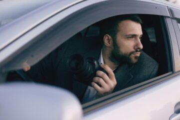Mężczyzna z brodą siedzi w samochodzie, wychylając się przez okno i trzymając kamerę, wyglądając na skupionego i czujnego, gdy bada możliwą zdradę żony.
