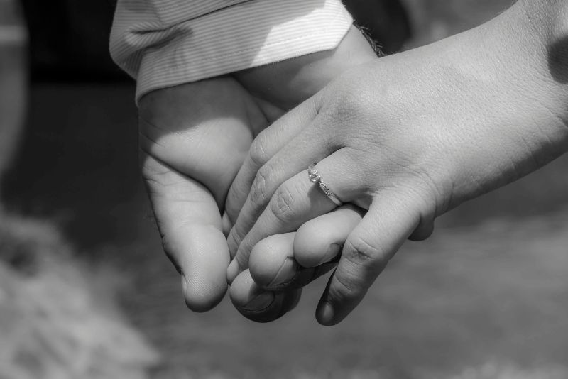 Zbliżenie dwóch osób trzymających się za ręce, jedna z nich nosi na palcu obrączkę, a ciągłe trzymanie partnera za rękę. Obraz jest czarno-biały z delikatnie rozmytym tłem.