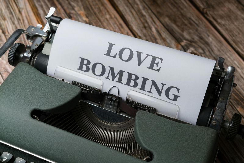 Maszyna do pisania z pogrubioną kartką papieru z napisem "LOVE BOMBING", ustawiona na rustykalnej drewnianej powierzchni - subtelny ukłon w stronę tej manipulacyjnej taktyki.
