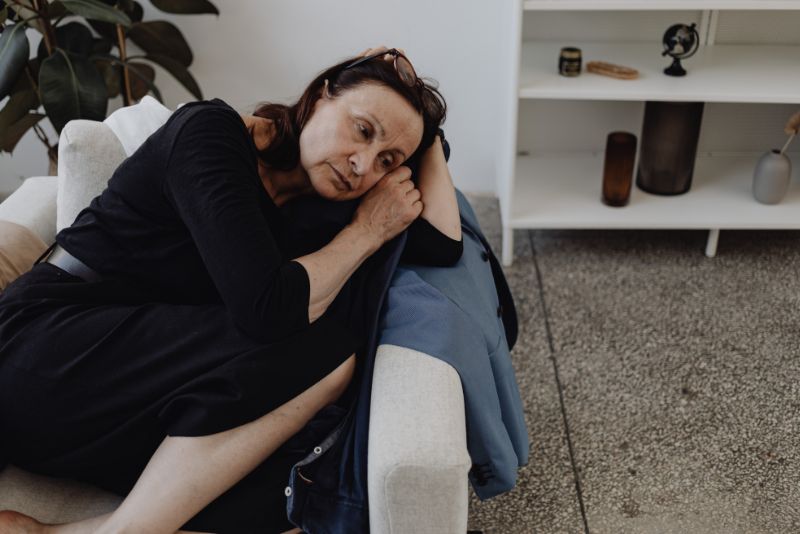 Kobieta w czarnej sukience siedzi skulona na jasnej kanapie, opierając głowę na ramieniu i wyglądając na zamyśloną lub smutną. W tle widoczna jest roślina i biały regał.