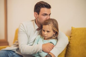 Mężczyzna siedzi na żółtej kanapie, przytulając mocno młodą dziewczynę. Oboje wyglądają na poważnych i zatroskanych - obraz odzwierciedlający parentyfikację, gdy dziewczyna z jego ramieniem owiniętym wokół jej ramion.
